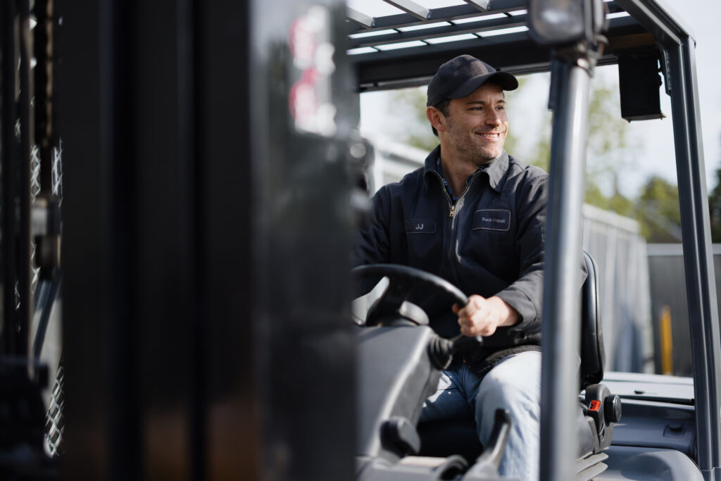 Lagerarbeiter, der lächelnd einen Gabelstapler fährt und dabei Fachwissen und Effizienz in einer geschäftigen Logistikumgebung unter Beweis stellt.